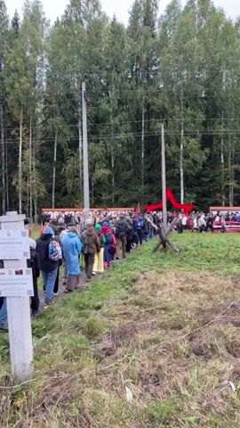 The video shows a procession of people walking along a path in a grassy, wooded area. The path is lined with tall trees. In the background, there are large banners with red lettering and images. A group of people are gathered at the end of the path, some standing near tractors and what appears to be a stage or platform. Red fabric, possibly a banner or decoration, is visible in the background. There are also power lines and poles along the path. The weather appears overcast. Several signs are visible on a wooden post in the foreground, with Russian text on them.