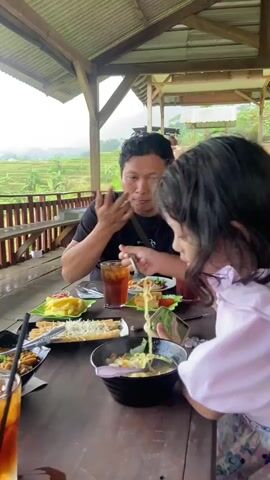 A young girl with long, dark hair is eating noodles out of a black bowl on a wooden table in an open-air restaurant.  She is wearing a light pink sweatshirt and is hunched over the bowl, bringing the noodles close to her face with a fork. To her left, an older man wearing a black t-shirt is sitting and eating at the same table. The table is covered with multiple plates of food, including spring rolls, french fries, and a dish with dark sauce. There are also glasses of iced tea on the table. Behind the man, a railing surrounds the restaurant’s edge, and in the background, a green rice paddy field is visible under an overcast sky. The restaurant structure has a wooden frame and a corrugated metal roof.