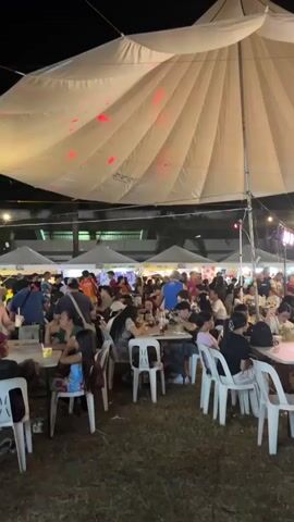This is a wide shot of an outdoor night market under a large white tent. The tent has blue and red lights projected onto it. The market is crowded with people sitting at tables and walking around. There are food stalls in the background. The overall atmosphere appears festive.