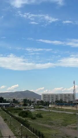 A small dark object, possibly an aircraft or drone, flies across a bright blue sky filled with scattered white clouds. Below the sky, a range of buildings, including apartment blocks and a tall telecommunications tower, are visible, situated against a backdrop of distant mountains. The scene appears to be an urban or semi-urban environment during daylight.