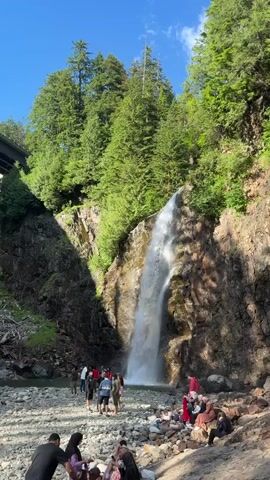 A scenic view captures a tall waterfall cascading down a rocky cliff face into a shallow riverbed. The area is surrounded by lush green trees and a clear blue sky. Several people are scattered around the riverbank, some standing near the waterfall and others sitting on the rocks. They appear to be enjoying the natural beauty of the location. There is a bridge structure visible in the upper left corner of the frame. The overall atmosphere suggests a peaceful and recreational outdoor setting.