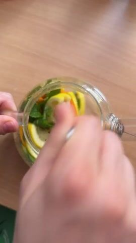 The video shows a close-up, overhead shot of a glass teapot filled with slices of orange and lemon, and sprigs of mint. The teapot is placed on a wooden table. Two hands are visible, one holding the handle of the teapot and the other stirring the contents with a spoon. The lighting suggests daytime, and there are no external environmental cues to indicate weather. The focus is on the preparation of a beverage, likely infused water or tea, with the stirring action creating a sense of movement and freshness. There is no visible text in the video. The scene is domestic and casual, suggesting a home environment. The activity is the mixing of ingredients for a drink.