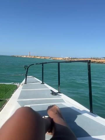The video shows a person lying on the bow of a boat, presumably enjoying a ride. The camera is positioned close to the person's lower legs and feet, looking forward. The boat is on the water, moving toward a shoreline with a lighthouse in the distance. The water is turquoise, and the sky is clear blue. Another sailboat passes by, full of people. The deck of the boat has some artificial turf on it.