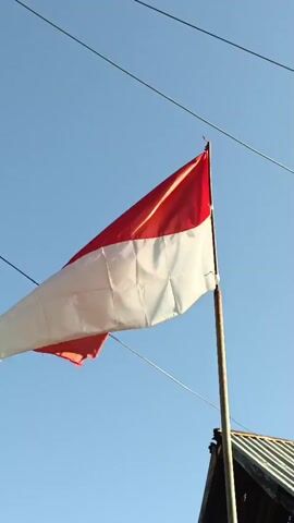 A red and white flag, likely the Indonesian flag, is attached to a metal pole and waves in the wind against a clear blue sky. The flag appears slightly torn in places. Two thin black wires are stretched across the upper portion of the frame. A portion of a roof, made of corrugated metal and wood, is visible in the lower right corner. The camera is positioned low, looking upwards at the flag.