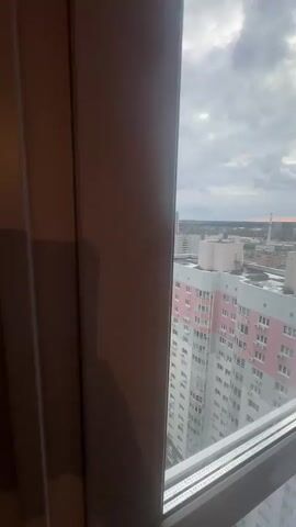 The video opens with a close-up shot of a white window frame, slightly ajar, with a brown vertical element on the left, possibly part of a curtain or another window frame. As the window is pushed open, a panoramic view of an urban landscape unfolds from a high vantage point.

The scene reveals numerous multi-story residential buildings under a cloudy, overcast sky. Many of the buildings are light-colored, with one prominent building in the foreground painted in shades of pink and white, featuring multiple air conditioning units on its facade. Beyond this building, a street with parked cars and some trees is visible. In the distance, more residential complexes, some with colorful facades (like light green, blue, and yellow), stretch across the horizon. A tall, slender structure, possibly a chimney or communication tower, is visible on the distant horizon to the right, where a faint orange glow suggests either sunrise or sunset. The overall impression is that of a bustling city in Edem, Russia, viewed from a high-rise apartment on an overcast day. No discernible text is visible in the distant cityscape or on the buildings.