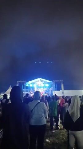 A crowd of people are gathered at night, facing a stage with bright lights. Smoke or fog is being used for lighting effects. A concert or performance is taking place on the stage.