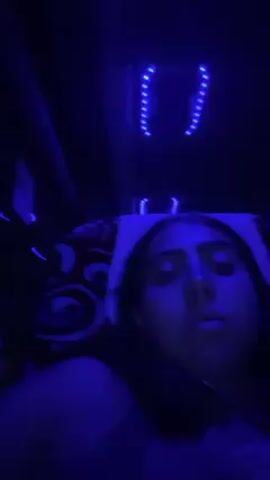 The video shows a young woman lying down, eating from a container with a spoon. The lighting is dark blue, and there are strings of blue lights visible on the ceiling above her. A pillow with a patterned cover is behind her head. She looks directly at the camera as she eats. The setting appears to be inside a vehicle, possibly a bus or a car.