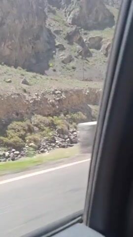 The view from a moving vehicle travels along a paved road next to a rocky hillside. The roadside is lined with concrete barriers. The hillside is mostly gray and brown with sparse green vegetation. The sky is bright with some clouds.