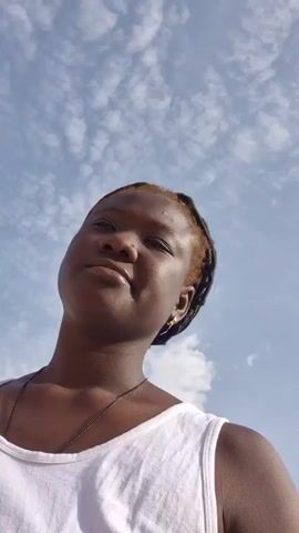 A woman is looking up at the sky. She is wearing a white tank top and a necklace. Her hair is styled in dreadlocks, with some strands dyed blonde. She has a nose ring and gold earrings. The sky is blue with scattered clouds. The camera is held at a low angle, looking up at her.