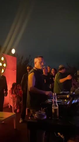 The video shows a crowded outdoor event at night. In the foreground, a person is operating DJ equipment, with a bottle of beer on the table. There are several other people visible in the background, some dancing and others watching. The lighting is dim, with spotlights creating beams in the darkness. The overall atmosphere appears to be a lively party or concert.