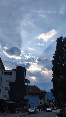 This media file captures a street view in Bolzano, Italy, during what appears to be late afternoon or early evening. The sky is a prominent feature, with a dynamic mix of blue sky and scattered clouds. Dramatic crepuscular rays, illuminated by the setting sun, pierce through the clouds, casting shadows on the buildings below.

In the foreground, to the left, a multi-story building with a dark facade is visible, featuring balconies. To its right, a lighter-colored building with multiple windows is partially obscured. A vibrant green tree stands tall on the right side of the frame, its leaves dense.

The street below is lined with several buildings of varying colors and architectural styles, including a blue building with a red-tiled roof. Several cars are parked on the street and a few are in motion, suggesting normal daytime activity. A person is seen on a bicycle near the left edge of the frame, further indicating everyday life.

The lighting suggests a transition from day to evening, with the sun's rays creating a dramatic and somewhat serene atmosphere. The presence of mountains in the distant background confirms the mountainous terrain characteristic of Bolzano. No text is visible in the media file.