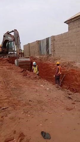 A Volvo excavator digs a trench in red soil near a half-finished building. Two workers in vests and hard hats work inside the trench, one kneeling and one standing.  A small group of people is visible near trucks in the background. The sky is overcast.