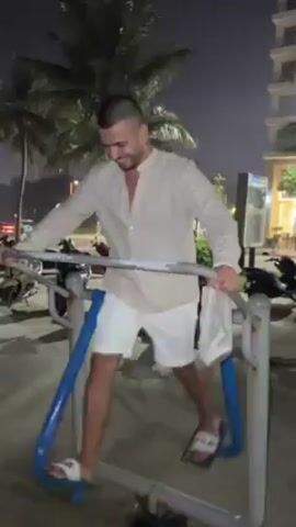 A medium-angle shot at night features a man using outdoor exercise equipment. The man is fair-skinned, has a shaved head, is wearing a light tan button-down shirt, white shorts, and white slide sandals. He is smiling, looking at the equipment as he moves his legs in a striding motion. The equipment is constructed of metal and is painted silver and blue. Parked motorbikes are visible in the background, as are tropical trees and a tall building. The scene is lit by artificial light.