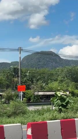 The video opens with a view of a large, lush green mountain under a bright blue sky with scattered white clouds. In the foreground, there are power lines crisscrossing the frame. A utility pole stands on the left side. A red and white road barrier is visible at the bottom of the frame. A gray vehicle passes by on the left. A bright orange sign with white Thai text is visible on the utility pole. More trees and vegetation fill the middle ground, and a simple structure with a white roof is also present. The camera pans slightly to the right.