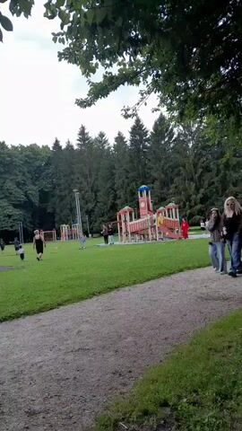 A group of people are in a park, with a playground in the background. Some are walking on a path, while others are on the grass. There are trees and a cloudy sky.
