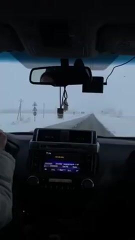 The video shows the view from inside a car driving on a road covered in snow. The sky is overcast. The car's dashboard and radio are visible, with the radio tuned to 101.80 MHz. The driver's hands are on the steering wheel. A passenger is in the passenger seat. A rearview mirror hangs from the ceiling. Another vehicle is ahead on the road. The environment appears cold and snowy.