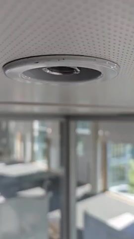 A close-up shot focuses on a circular ceiling vent with a metallic ring. The background is blurred, showing a large window or glass partition with reflections of an office environment, including desks and chairs. The lighting appears to be natural, coming from the window. The overall scene suggests an indoor setting, likely an office or a modern building.