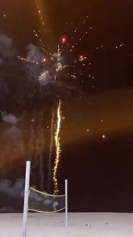 The video shows a nighttime scene on a sandy beach with a volleyball net in the foreground. Fireworks are being set off, creating bursts of light and smoke in the dark sky. The fireworks vary in color and intensity, with some appearing as bright sparks and others as larger explosions. The beach appears empty except for the volleyball net and scattered debris, possibly from previous fireworks. The lighting suggests the presence of artificial light sources in the distance, possibly from buildings or streetlights, casting an orange glow on the horizon. The sound of fireworks is audible throughout the video.