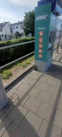 The video shows a green ticket machine at a bus stop. The machine has buttons for tickets, S, U, and a bus. There is a small green bottle on the ground next to the ticket machine. The ground is paved with gray and tan interlocking pavers. There is a metal fence to the left of the ticket machine, and behind the fence is a grassy area with some bushes and trees. In the background, there are houses and a blue sky with some clouds. The video pans slightly to the right, revealing more of the ticket machine and the bus stop shelter.