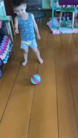 A young child is playing with a ball on a wooden floor indoors. The child, wearing a blue tank top and light blue shorts, is barefoot. A multi-colored ball with green and purple stripes is on the floor in front of the child. The room appears to be a home with wooden floorboards and furniture visible in the background. A colorful blanket or rug is draped over a piece of furniture on the left. There is some text visible on the ball, which is too blurry to read. The child's posture and movement suggest they are actively engaged in playing with the ball, possibly kicking or rolling it. The lighting suggests it could be daytime.