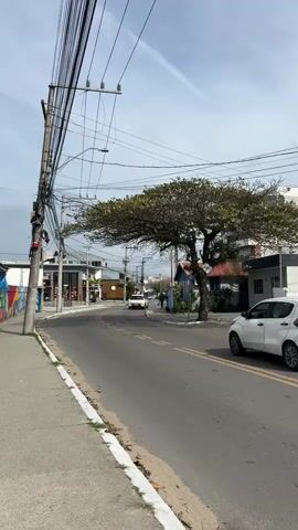 A white hatchback drives down a paved road. To the left of the road is a sidewalk with a white painted curb, and a utility pole with many wires. To the right of the road is a tree with green leaves and houses. The sky is blue with some clouds. The camera is positioned on the sidewalk to the left of the road, looking forward.