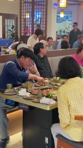 A group of people are dining at a Korean BBQ restaurant. There are two tables visible. On the closest table, a man in a blue shirt is cooking meat on the grill, using chopsticks to arrange it. Other people sit at the table with him, including a woman in a yellow checkered shirt. The table is covered with small dishes of food. The second table has more people at it. The restaurant has hanging lantern decorations, and other patrons can be seen in the background.