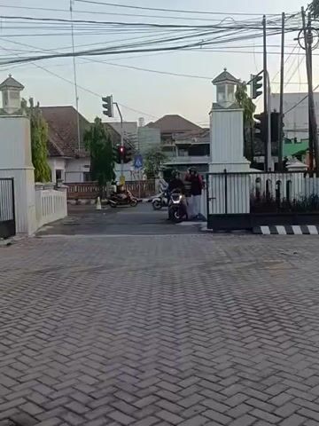 The video shows a street scene with a paved walkway in the foreground. In the middle ground, there is a road with a traffic light that is red. Two people are standing next to a motorcycle, which is parked on the road. There are buildings and utility poles in the background, and the sky is visible above. The overall lighting suggests it is either early morning or late afternoon.