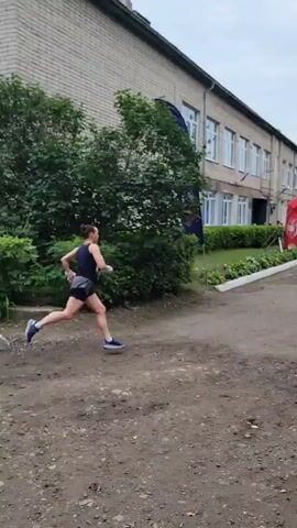 A woman in athletic wear runs towards an inflatable archway marked with Russian text that translates to "FINISH." Several other people are gathered behind the arch, some appearing to be participants in a race or event, while others may be spectators or organizers. The environment suggests an outdoor setting, possibly a town or school area, with a building in the background and trees to the left. The ground appears to be a mix of dirt and gravel. The lighting indicates it is daytime, with an overcast sky. The woman is in the foreground, running away from the camera, with the finish line arch in the midground.