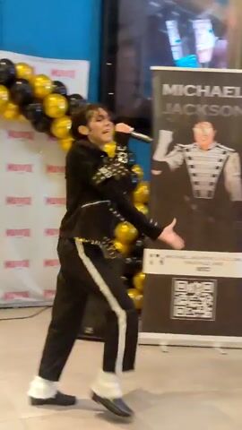 A male impersonator dressed as Michael Jackson, wearing a black sequined jacket with white accents, black pants with a white stripe, and white fur cuffs on his shoes, performs in front of a banner featuring a picture of Michael Jackson. The impersonator is holding a microphone and appears to be singing and dancing. He is positioned in the center of the frame, with his body turned to the left and his right arm extended. Behind him, the banner has the text "MICHAEL JACKSON" at the top. The scene is decorated with black and gold balloons arranged in arches on either side of the banner. To the left of the impersonator, there is a dark speaker. The background wall is blue, and there are other banners visible to the right, partially obscured. The lighting suggests an indoor setting, possibly a promotional event or performance space. The impersonator's facial features and hairstyle resemble Michael Jackson's.