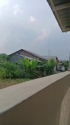 The video captures a view from a balcony looking out at a cloudy sky with visible raindrops. In the distance, there are houses surrounded by lush green vegetation. A person's hand is visible on the right side of the frame, seemingly reaching out from the balcony. The focus is on the rainy atmosphere and the outdoor environment.
