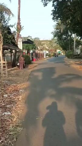 The video shows a paved road in what appears to be a rural or suburban area. The sun is casting long shadows across the road, indicating it is either early morning or late afternoon. On the left side of the road, there is a small structure with a thatched roof, possibly a stall or a small building, with some wooden supports. Two women are standing near this structure, one facing away from the camera and the other facing towards the camera. The woman facing the camera is wearing a light-colored top and light-colored pants, and she has a headscarf. The other woman is wearing a darker top and pants. Behind them, there is a fence or wall decorated with red and black ornamental posts. Further down the road, there are trees and more structures, including what looks like a building with a white facade and some greenery. A single vehicle, possibly a motorcycle, is visible in the distance on the road. The ground along the side of the road is covered with dry leaves and some grass. The overall impression is a peaceful, sun-drenched scene.