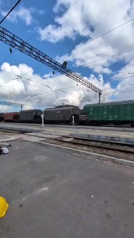 The video pans across a train station platform. In the foreground, on the asphalt, are several yellow, dome-shaped bollards and a collection of bags and hard hats. A black dog lies on the platform near the tracks. In the background, a long freight train is stopped on the tracks, consisting of several green and grey hopper cars. Overhead, electrical lines for the train are visible against a cloudy blue sky. The camera moves slowly from left to right, revealing more of the train and the station environment.