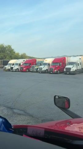 The video shows a row of parked semi-trucks at what appears to be a truck stop or rest area. The camera is positioned from inside another semi-truck, looking forward. The sky is clear and blue, with the sun setting or rising, casting a soft light on the scene. The trucks are of various colors, including red, white, and blue. Some trucks have trailers attached. A GPS device is visible on the dashboard of the camera's truck, displaying a map. The ground is paved asphalt. In the distance, there are trees and mountains. The video appears to be a static shot with no movement.