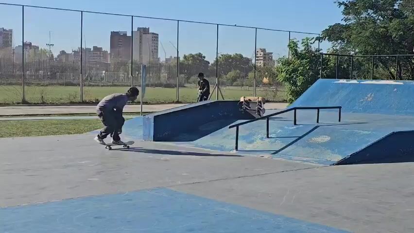 The video shows a skateboarder in a skate park. The skateboarder is wearing a gray shirt and black pants. He is skating on a blue ramp with a black rail. In the background there are other skaters, a chainlink fence, a field and some buildings. The video is taken outdoors on a sunny day.