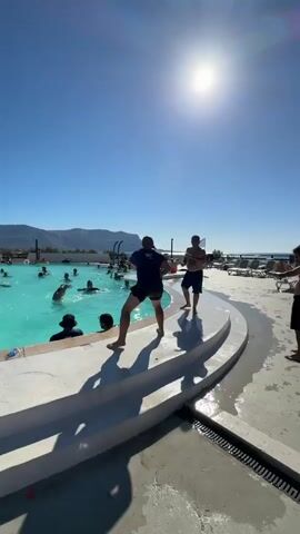A group of people are gathered around a large, blue swimming pool on a sunny day. Two men are standing at the edge of the pool, facing each other, and appear to be in the middle of a game or activity, possibly a water fight, as one of them is holding what looks like a water gun. Several other people are in the pool, splashing and interacting. Deck chairs are visible in the background, suggesting this is a resort or public swimming area. The sky is clear and blue, with the sun high overhead, casting long shadows on the ground. The overall atmosphere is one of leisure and recreation.