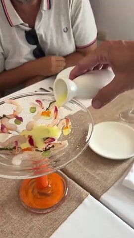 A close-up shot shows a person's hand pouring a pale green sauce from a white pitcher onto a clear glass dish filled with food. The dish is elevated on an orange, swirled glass base. The food in the dish appears to be thinly sliced raw shrimp, garnished with small red spheres (possibly roe), green elements, and purple petals. A man in a white polo shirt is visible in the background, seated at a table covered with a neutral-colored tablecloth. The table setting includes white plates and a glass of amber liquid. The lighting is bright, suggesting an indoor or shaded outdoor setting.