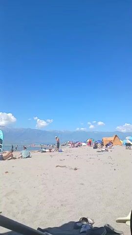 The video captures a sunny day at a crowded beach. The sand is light brown, and the sky is a clear, bright blue with a few white clouds scattered near the horizon. In the distance, a mountain range with snow-capped peaks stretches across the background. Numerous people are spread across the beach, some lying on towels, others standing or walking. Several colorful tents and umbrellas provide shade for beachgoers. A stand-up paddleboard stands upright on the sand near the left side of the frame. In the foreground, a dark shadow obscures part of the view, with a pair of shoes and other objects partially visible. The water is visible in the middle ground, with people swimming and wading. The overall scene depicts a lively and enjoyable beach atmosphere.