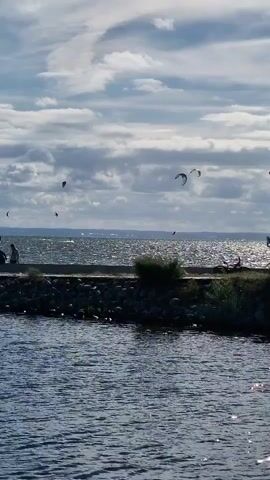 The video shows a wide shot of a body of water with a cloudy sky. Several kite surfers are visible on the water, appearing to be in motion. A breakwater made of rocks separates the foreground water from the body of water where the kite surfers are. There are a few people on the breakwater. The sun is shining, creating a shimmering effect on the water. The overall scene depicts a recreational activity on a partially cloudy day.