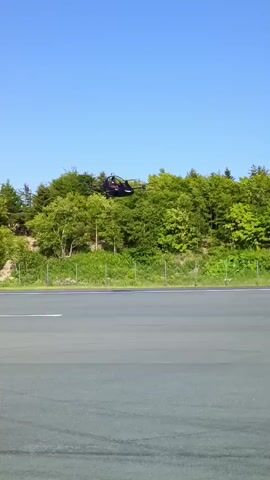The video shows a black drone with four propellers and a cabin flying in the air. The drone is moving from left to right across the screen. It is flying over a grey paved area with some tire marks. Behind the paved area is a line of green trees. The sky is clear and blue. The drone is in focus and the background is slightly blurred. The drone appears to be a prototype for a personal flying vehicle.
