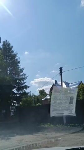 This video captures a view from inside a vehicle as it passes by a light blue and white Orthodox church in Karpovo, Russia. The scene appears to be set on a bright, sunny day, with the sun creating lens flares and bright light illuminating the sky and the trees. The church is a prominent subject, with its distinctive domes topped with crosses. There are also other structures visible, including a fence and a small red brick archway.

A yellow triangular road sign with a red border is visible on the left side of the screen. The sign depicts a pedestrian crossing and also has the number "50" in a red circle, indicating a speed limit. There is also text on the sign, though it is partially obscured and appears to be in Cyrillic.

A piece of paper with printed text and a QR code is attached to the inside of the vehicle's windshield, obstructing the view slightly. The paper appears to be a document or a notice of some kind. The camera is positioned low, suggesting it is looking out from the dashboard or lower part of the windshield.

The overall impression is that of a drive through a village or town on a clear day, passing by a significant religious building. The presence of the speed limit sign and pedestrian crossing suggests the vehicle is on a road with potential traffic and pedestrian activity. The clear blue sky and bright sunlight indicate fair weather, likely during the daytime.