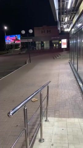 The video shows a street scene at night. A paved walkway leads down a slight slope from right to left. A metal railing runs along the right side of the walkway. Beyond the walkway, there is a parking lot with visible parking space lines. Across the parking lot, there are several buildings. One building appears to be a store with "ФРУКТЫ" (Fruits) written on it. There are illuminated billboards and streetlights visible in the distance, indicating an urban or suburban setting. The overall atmosphere is quiet and dark, suggesting it is late at night or early morning. There are no people visible in the immediate foreground or midground, though there are parked electric scooters visible in the distance.