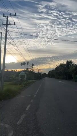 The video shows a paved road with white dashed lines in the foreground. On the left side of the road, there is a grassy field with some trees and buildings in the distance. A utility pole with many wires is also visible on the left. The sky is partly cloudy with the sun low on the horizon, suggesting it is either sunrise or sunset. The overall scene is rural and peaceful. There are no people or animals visible in the video.
