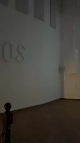 The video shows the interior of a dimly lit room, seemingly an art installation or gallery. The word "protos" is visible on the wall, seemingly embossed or made of a light material. Further into the room, there's an art piece featuring outlines of bottles. Towards the end of the clip, two figures, a woman and a man, are seen walking towards the artwork. Their figures appear mostly as silhouettes against the bright background. The overall environment suggests a contemporary or modern art space.