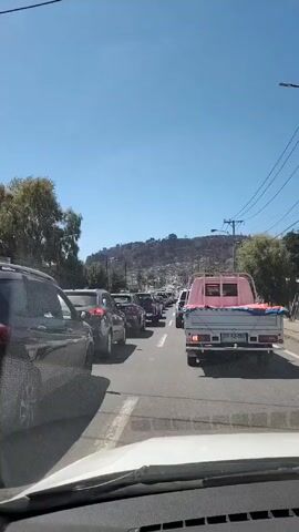 The video shows a congested two-lane road with cars lined up bumper-to-bumper. The view is from inside a car, looking out through the windshield.  A light blue pickup truck with a pink frame is directly ahead, and other cars are visible in both lanes.  A hillside covered with buildings is visible in the background under a clear, blue sky. Power lines run along the side of the road.