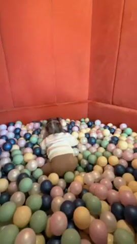 A toddler with pigtails is playing in a ball pit filled with a variety of pastel-colored balls. The ball pit has red upholstered walls. The toddler is wearing a white shirt and brown pants. She is crawling and playing with the balls. The camera is initially focused on the toddler, then pans up to show a woman holding the camera. The woman is wearing a brown top and has short blonde hair. She is smiling.