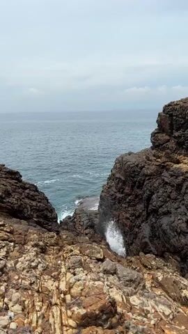 The video shows a rocky coastal landscape. The sea is visible in the distance under a cloudy sky. The camera is positioned above a rugged, rocky terrain, with large rock formations on either side, forming a narrow opening that leads to the sea. Waves crash against the rocks below, creating splashes of white foam. The overall environment is natural and somewhat barren, with the focus on the interaction between the sea and the rocky shore.