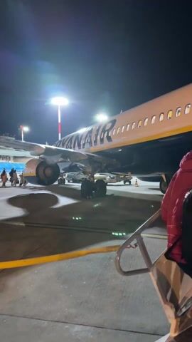 At night, a low-angle shot shows a Ryanair plane illuminated by bright lights. The camera follows two people walking up the portable stairs to board the plane. The first person is wearing a red jacket and a black backpack, while the second person is wearing a white puffer jacket. The airport tarmac is visible, along with support vehicles around the plane.