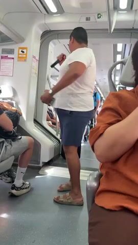 The video shows the interior of a train carriage, likely in Torremolinos, Spain, given the context. The primary subjects are several people, some seated and others standing. A man in a white t-shirt and blue shorts is the focal point, standing in the aisle and appearing to interact with something above him. He is holding a long, thin object, possibly a stick or a cane. To his left, another man is seated, wearing a black t-shirt and shorts. In the foreground, the arm and shoulder of a person wearing an orange shirt are visible, indicating they are also seated on the train.

The setting is a modern train carriage with white walls and fluorescent lighting. The floor is a light grey material. The doors between carriages are open, revealing more passengers and seating further down the train. There are luggage racks above the seats, and various signs are visible on the walls. One sign, in pink and white, has some text that is partially obscured, but "AVIS" and a symbol of a person walking are discernible. Another sign, yellow with black text and an exclamation mark within a triangle, is also visible.

The activity appears to be a performance or entertainment. The man in the white shirt is holding the object with both hands and gesturing upwards, as if he is juggling or performing some kind of magic trick. The other passengers seem to be observing him. The overall mood appears casual and possibly amused, as one might expect on public transport entertainment. The lighting suggests it is daytime, but there are no clear indications of the weather or specific time beyond that. The presence of a straw hat on top of a black speaker or bag in the aisle near the standing man suggests a potential performance setup.