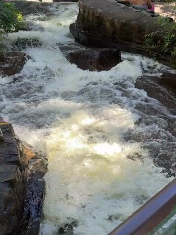 The video shows a whitewater creek cascading down a rocky bed. The creek flows from top to bottom of the frame, and there are several large rocks within its channel that cause the water to churn and foam. The surrounding environment includes green trees and vegetation on either side of the creek. On the right, there is a woman lying down on a rock, seemingly enjoying the scenery. A green metal railing is visible on the left, suggesting a viewing platform or walkway. The shot is taken from an elevated position, looking down at the flowing water and the surrounding area.