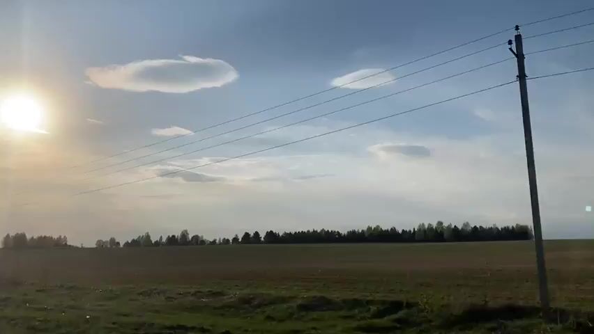 The video depicts a rural landscape on a sunny day. A large, open field with some green patches is visible in the foreground.  A line of trees marks the horizon. Power lines stretch across the sky, intersected by utility poles. The sun is bright and partly obscured by clouds. The camera appears to be moving forward.